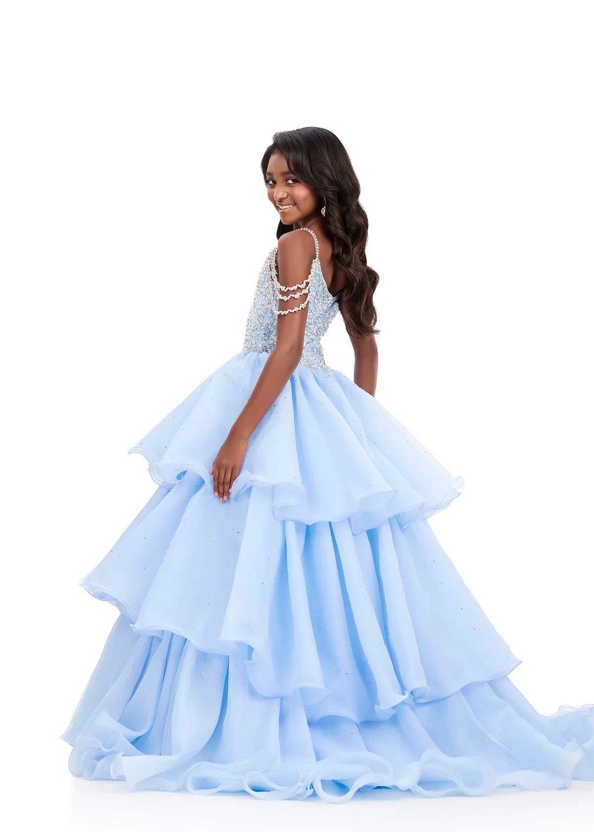 A young girl modeling a light blue quinceañera dress with a beaded bodice and a full, tiered tulle skirt, photographed against a solid white background.