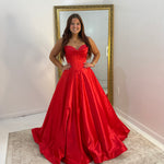 A woman wearing a bright red strapless ball gown with a sweetheart neckline and full skirt, standing in front of a gold-framed mirror in a room with white walls and a beige rug.
