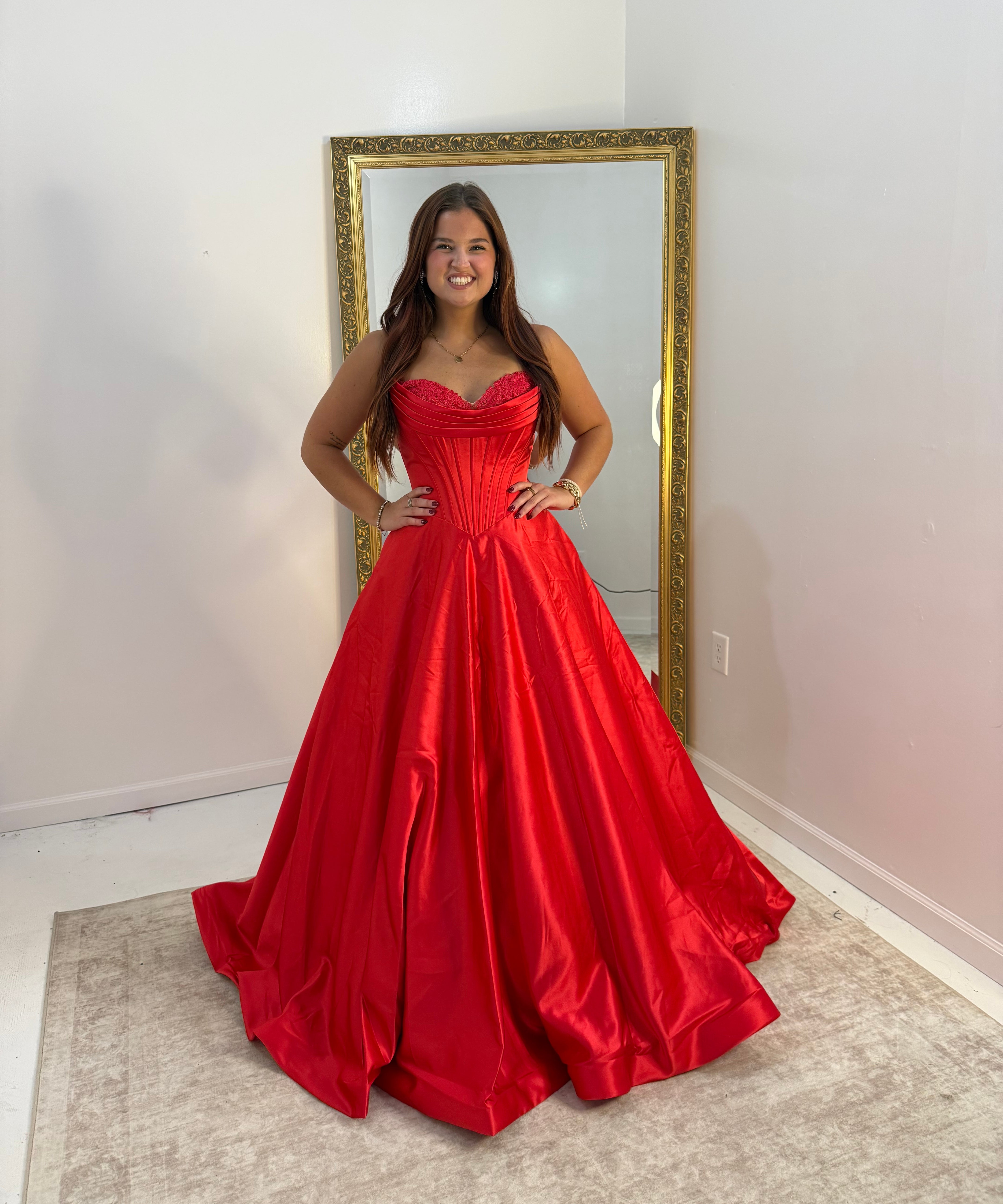 A woman wearing a bright red strapless ball gown with a sweetheart neckline and full skirt, standing in front of a gold-framed mirror in a room with white walls and a beige rug.