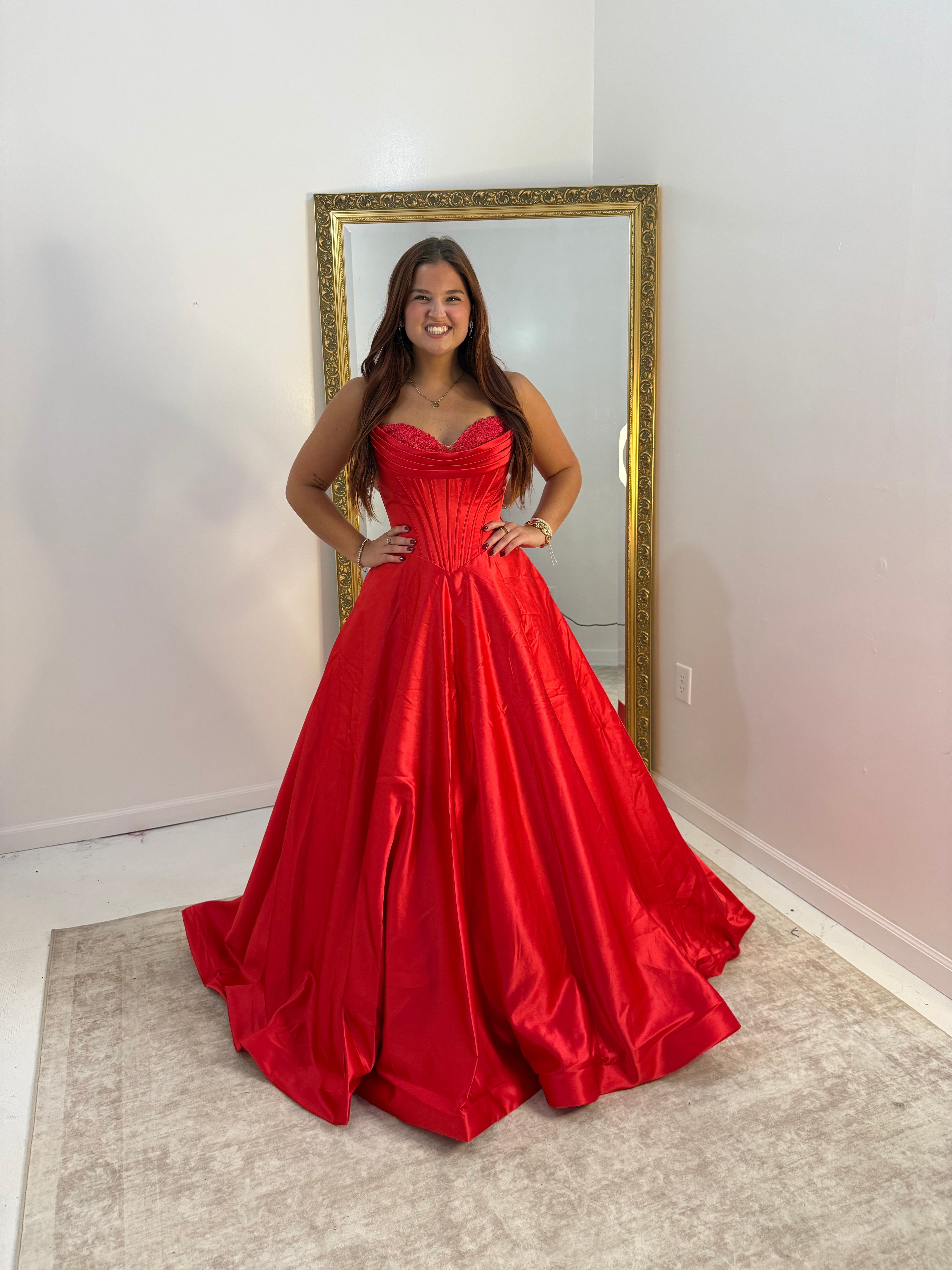 A woman wearing a bright red strapless ball gown with a sweetheart neckline and full skirt, standing in front of a gold-framed mirror in a room with white walls and a beige rug.