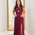 A woman modeling a sleeveless, floor-length burgundy maxi dress with a high thigh slit and a cutout at the chest, standing in front of a large gold-framed mirror in a white room.