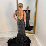A woman stands with her back to a large gold-framed mirror, wearing a black evening gown covered in silver rhinestone embellishments. The dress features a deep V-back, thin shoulder straps with black fabric accents, and a long train on a light-colored rug in a minimalist room.
