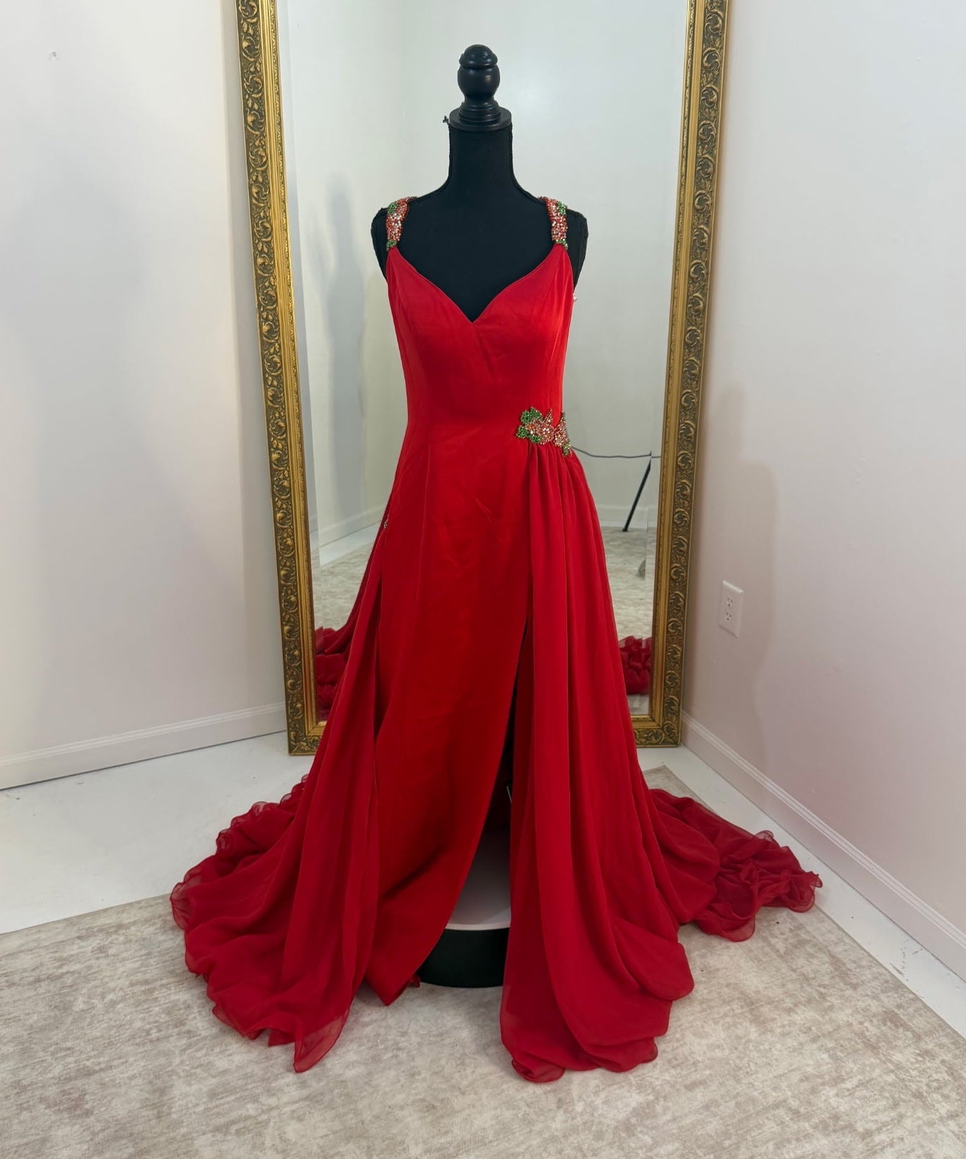 A bright red floor-length evening gown displayed on a black mannequin in front of a large gold-framed mirror. The dress has thin straps, a fitted bodice, a draped skirt, and a train spread across a light beige rug in a white room.