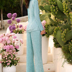 A woman stands outdoors on stone steps wearing a light aqua ribbed knit matching set consisting of a long-sleeve top and wide-leg pants. The scene is decorated with potted pink flowers and green cacti, and the brand name AMARRA appears in the bottom right corner.