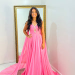 A woman wearing a bright pink floor-length formal gown with a sweetheart neckline and a long flowing train, standing indoors in front of a large gold-framed mirror.
