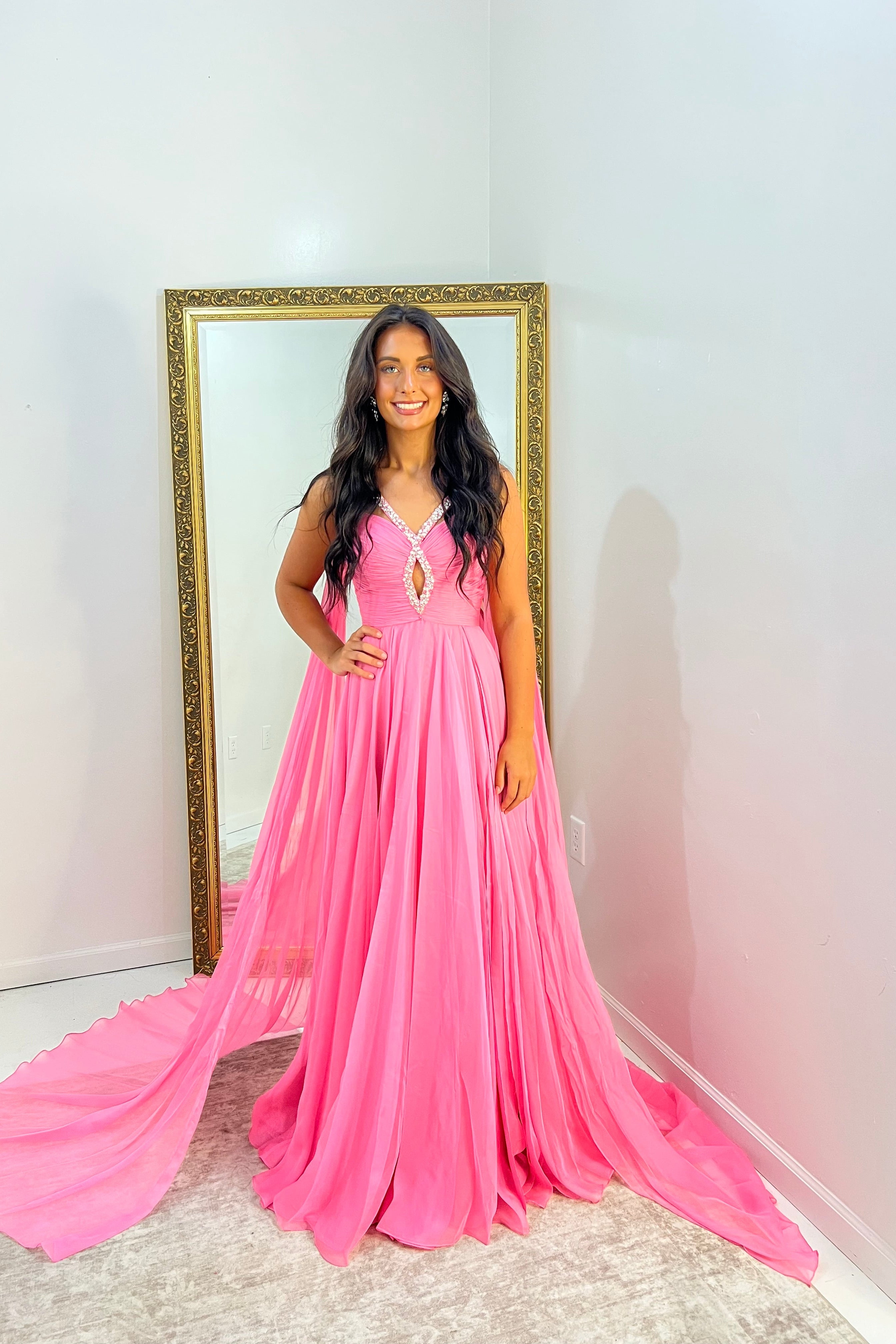 A woman wearing a bright pink floor-length formal gown with a sweetheart neckline and a long flowing train, standing indoors in front of a large gold-framed mirror.