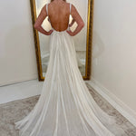 A woman stands with her back to the camera wearing a white dress with a low open back and long train, photographed indoors in front of a large gold-framed mirror.