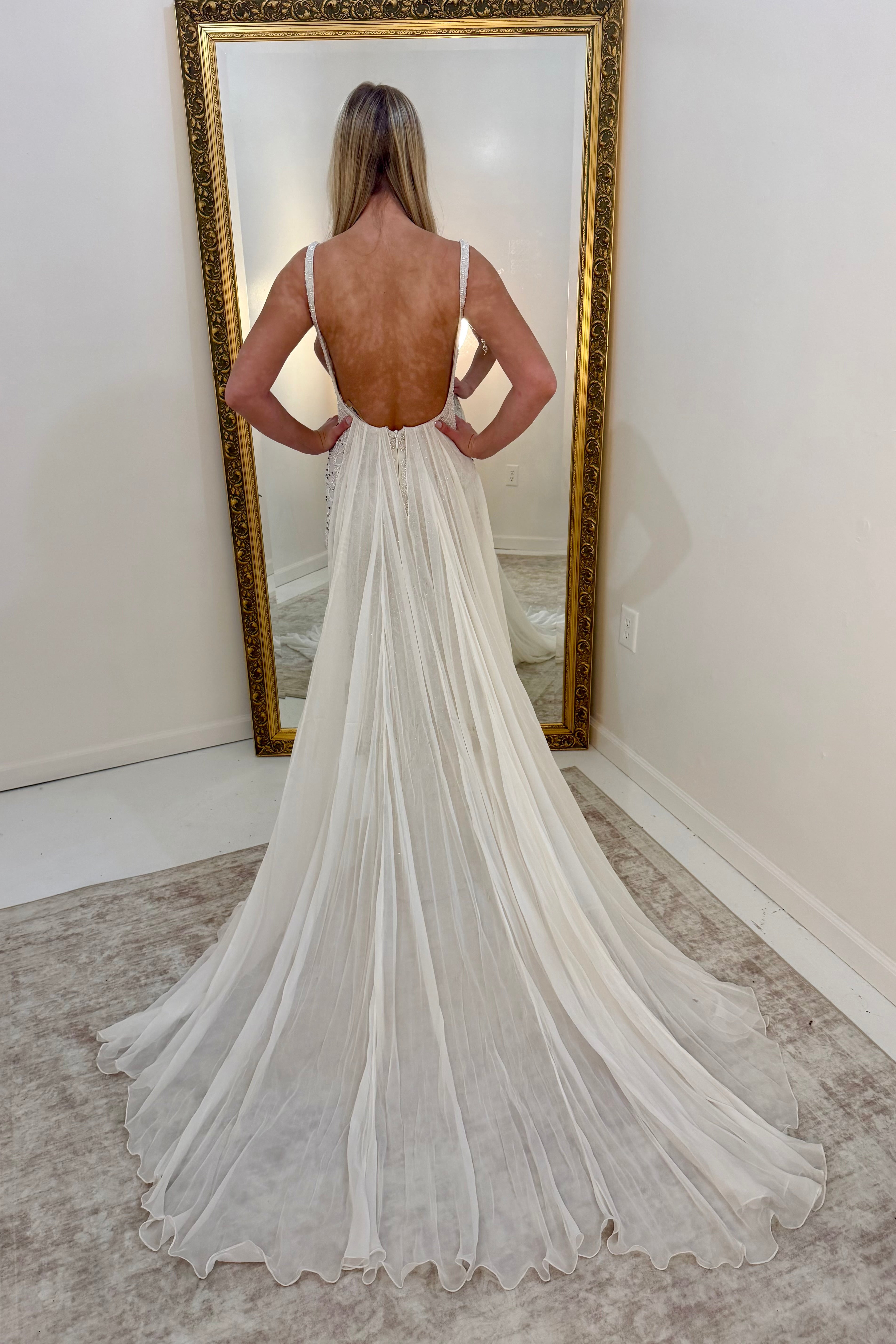 A woman stands with her back to the camera wearing a white dress with a low open back and long train, photographed indoors in front of a large gold-framed mirror.