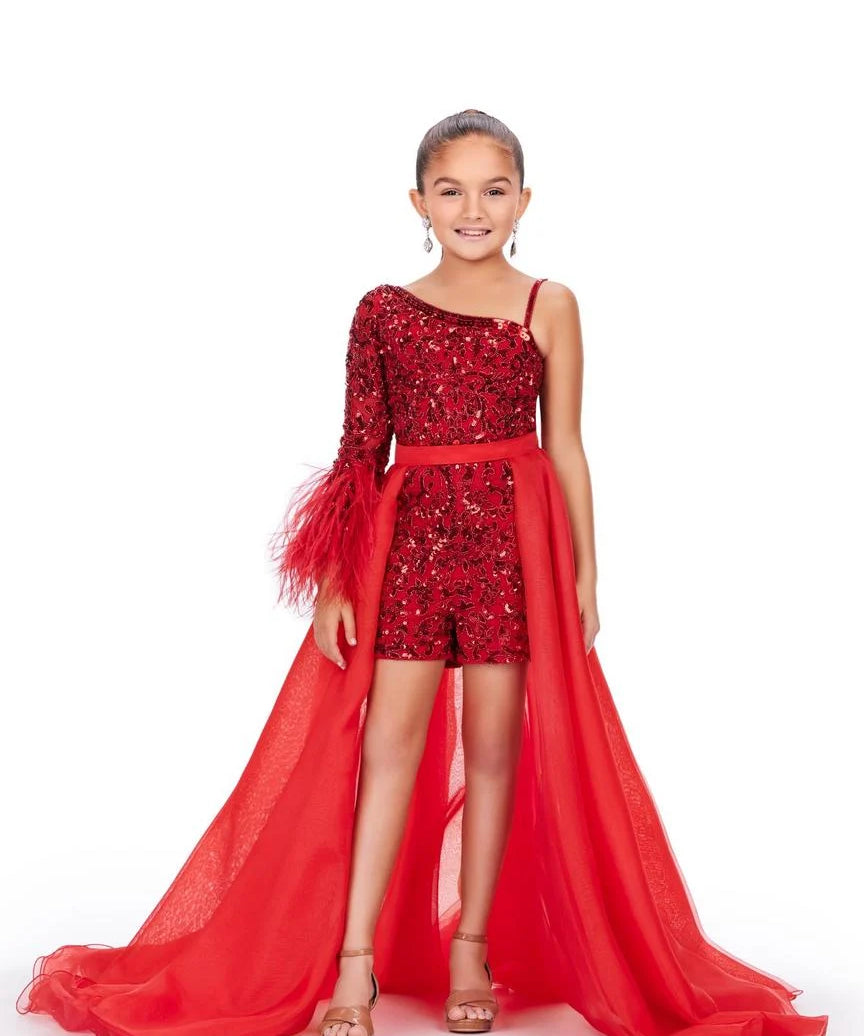 A young girl models a red organza overskirt atop a one shoulder beaded romper in front of a white background.