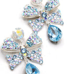 A pair of silver-tone drop earrings featuring a bow-shaped design and clusters of iridescent rhinestones, with light blue teardrop crystals, shown on a plain white background.