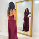 A woman stands with her back to the camera wearing a sleeveless, backless burgundy maxi dress, looking at her reflection in a large gold-framed mirror.