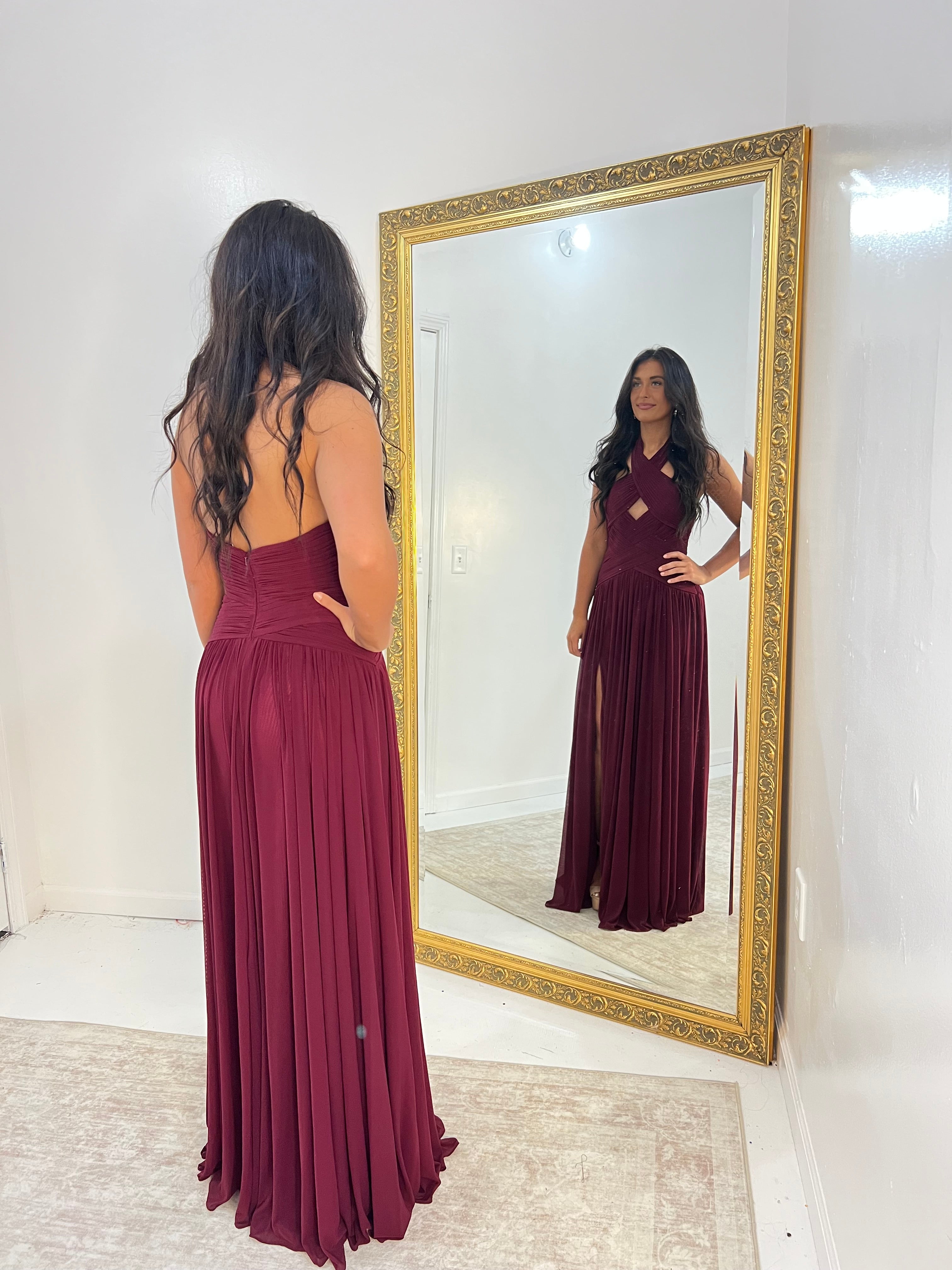 A woman stands with her back to the camera wearing a sleeveless, backless burgundy maxi dress, looking at her reflection in a large gold-framed mirror.
