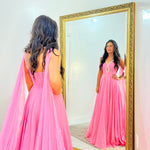 A woman wearing a bright pink floor-length gown stands in front of a large gold-framed mirror, looking at her reflection in a white room.