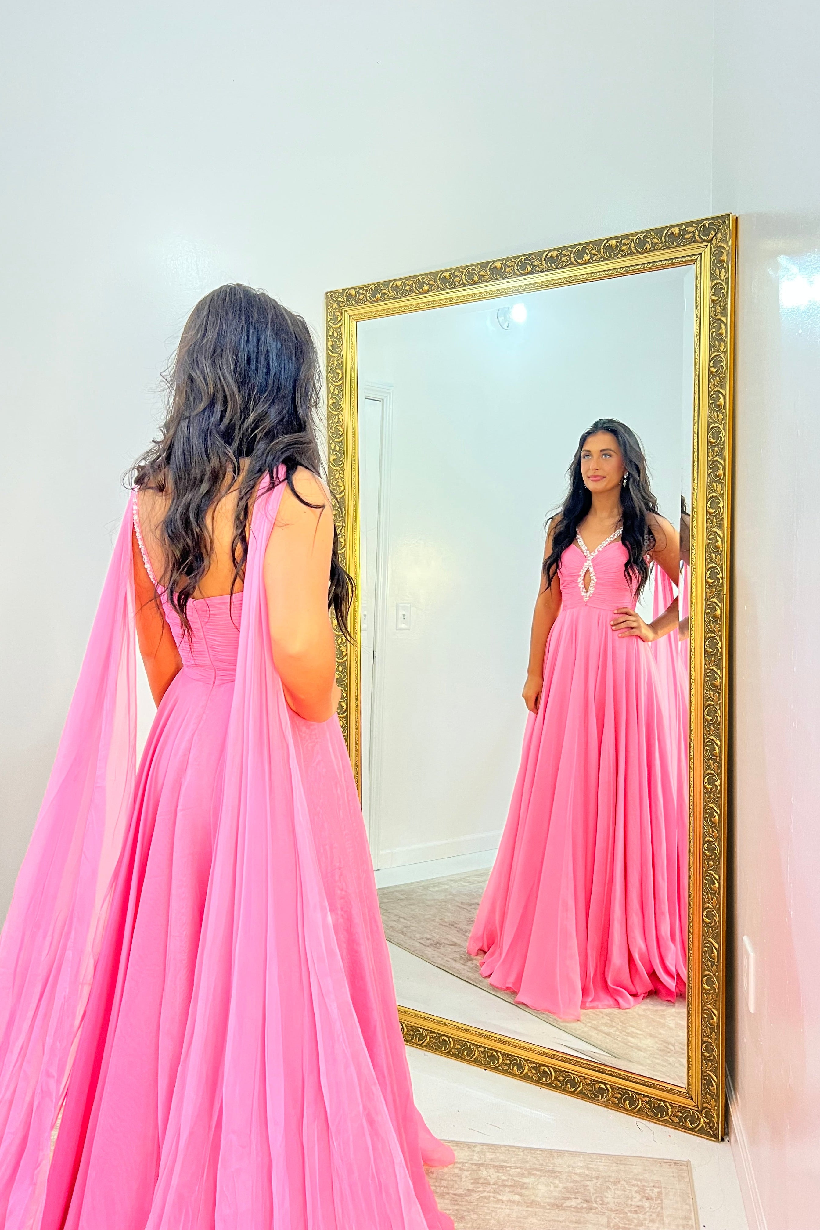 A woman wearing a bright pink floor-length gown stands in front of a large gold-framed mirror, looking at her reflection in a white room.