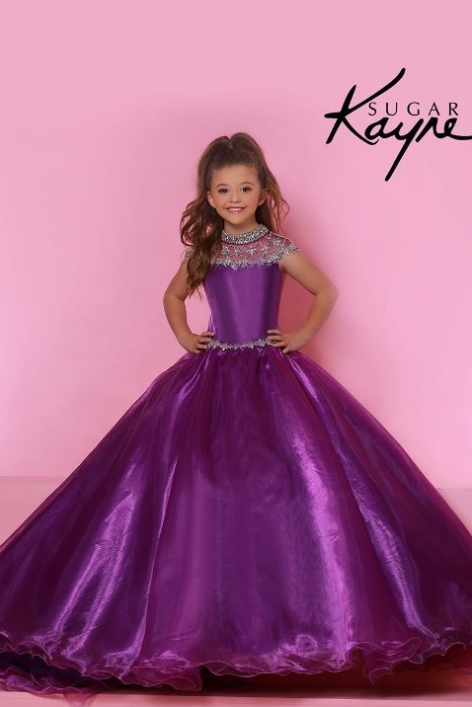 A young girl modeling a voluminous purple ball gown with a beaded high neckline and waistband, standing against a pink studio backdrop with the text 'SUGAR Kayne' in the upper right.