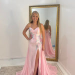 A woman stands indoors wearing a strapless light pink evening gown with floral embroidery, a thigh-high slit, and a long train. She is posed in front of a large gold-framed mirror that reflects her back and the full gown.