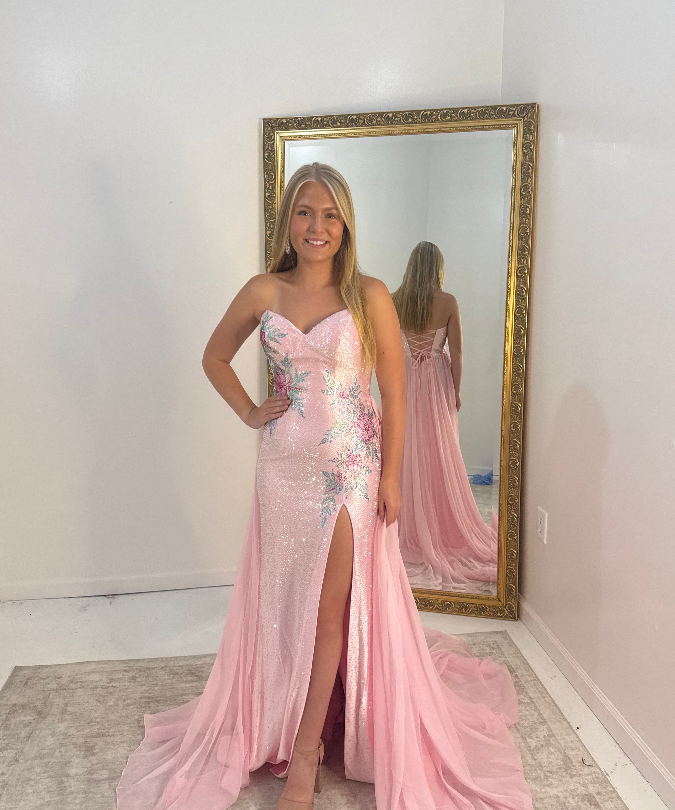 A woman stands indoors wearing a strapless light pink evening gown with floral embroidery, a thigh-high slit, and a long train. She is posed in front of a large gold-framed mirror that reflects her back and the full gown.