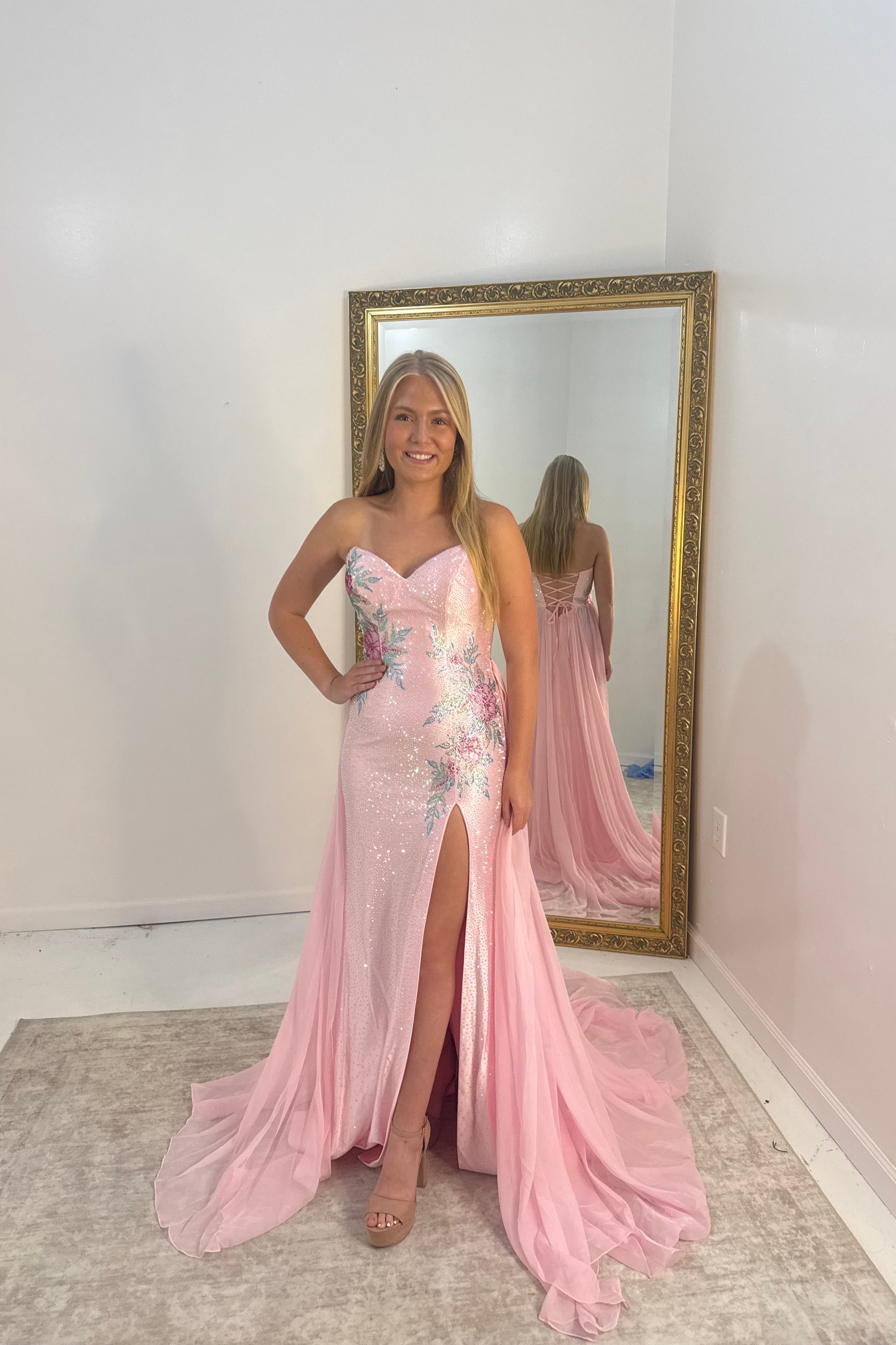 A woman stands indoors wearing a strapless light pink evening gown with floral embroidery, a thigh-high slit, and a long train. She is posed in front of a large gold-framed mirror that reflects her back and the full gown.