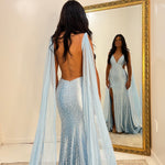 A woman stands with her back to the camera wearing a sparkling light blue mermaid gown with a long sheer cape train, standing in front of a large gold-framed mirror that shows her reflection.