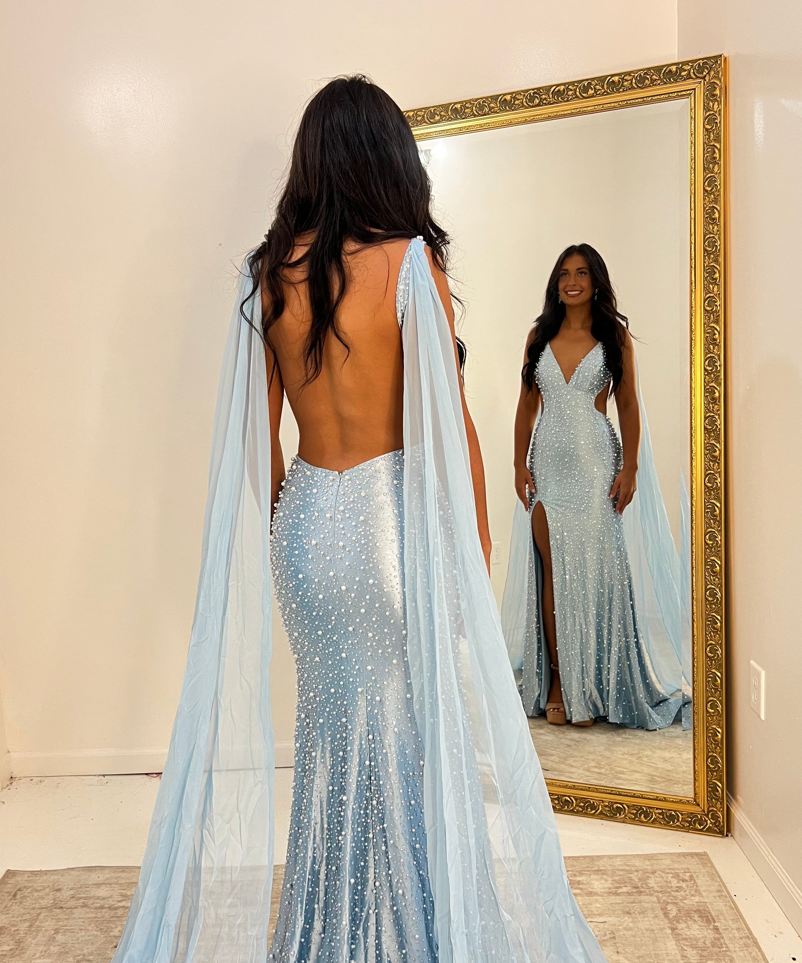 A woman stands with her back to the camera wearing a sparkling light blue mermaid gown with a long sheer cape train, standing in front of a large gold-framed mirror that shows her reflection.