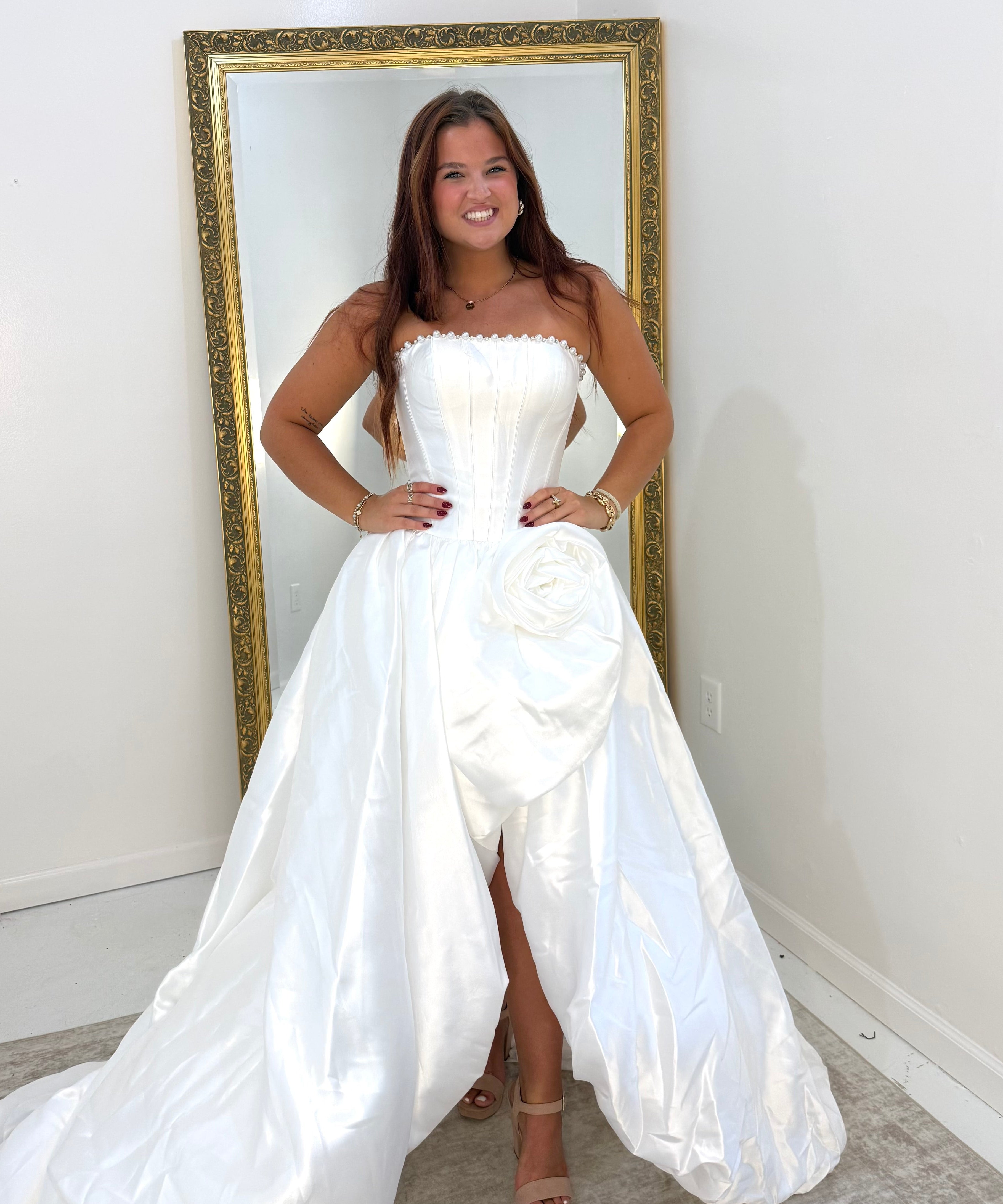 A woman wearing a strapless white satin gown with a structured bodice, large waist rosette, and dramatic overskirt/train, standing indoors in front of a gold-framed mirror.