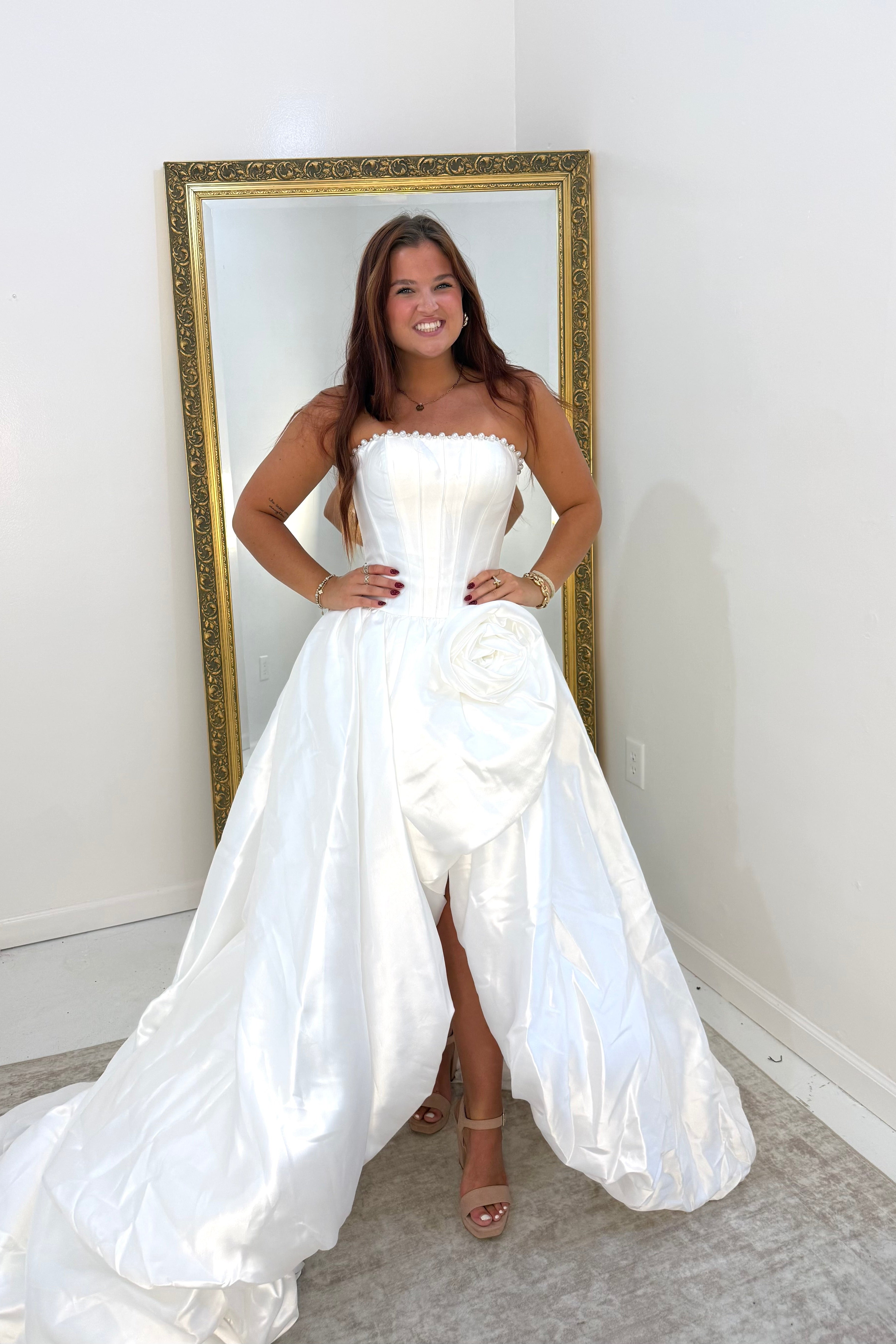 A woman wearing a strapless white satin gown with a structured bodice, large waist rosette, and dramatic overskirt/train, standing indoors in front of a gold-framed mirror.