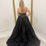 A woman stands with her back to the camera wearing a strapless black ball gown with a sheer lace bodice and a voluminous tulle skirt, posing in front of a large gold-framed mirror.