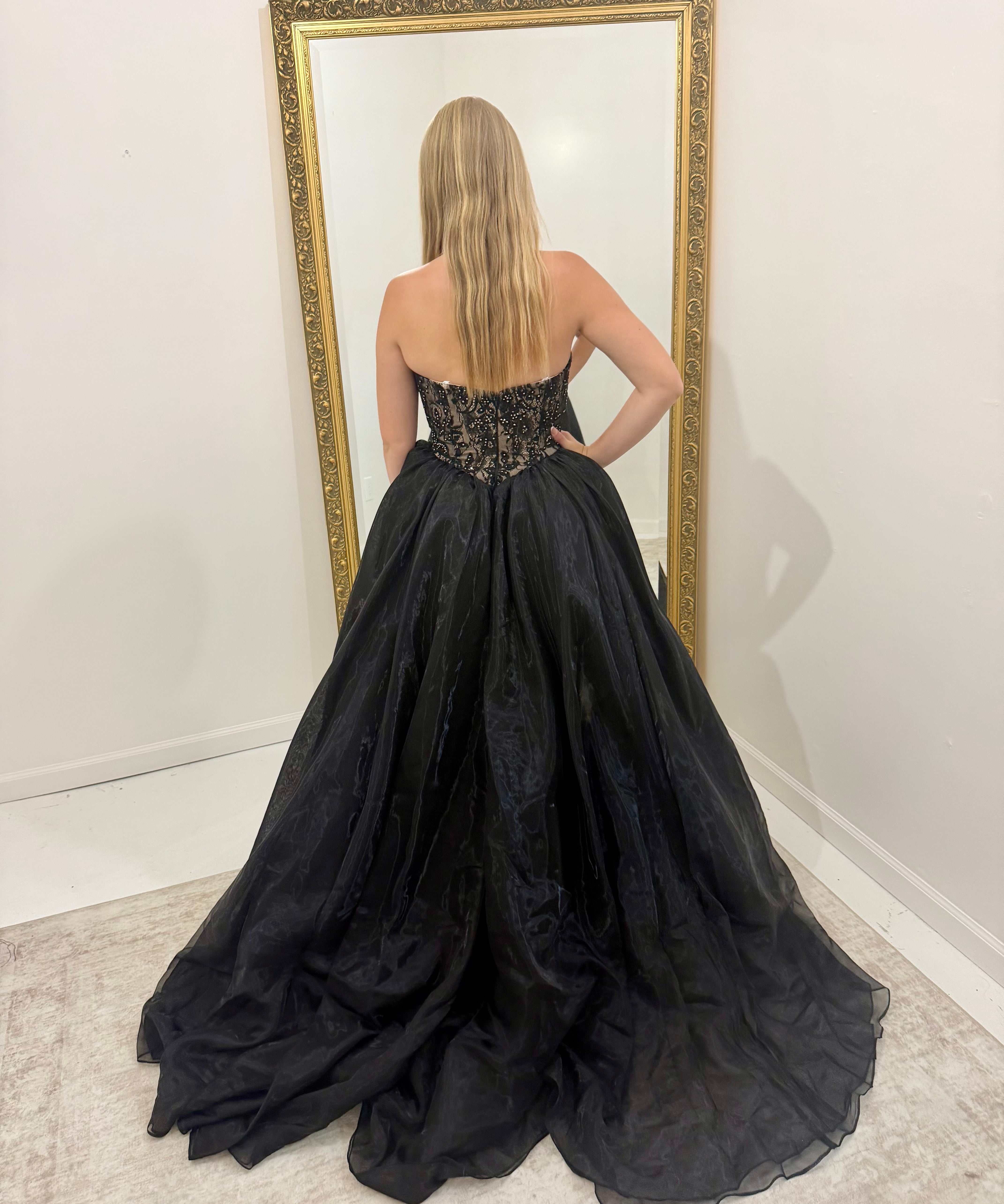 A woman stands with her back to the camera wearing a strapless black ball gown with a sheer lace bodice and a voluminous tulle skirt, posing in front of a large gold-framed mirror.