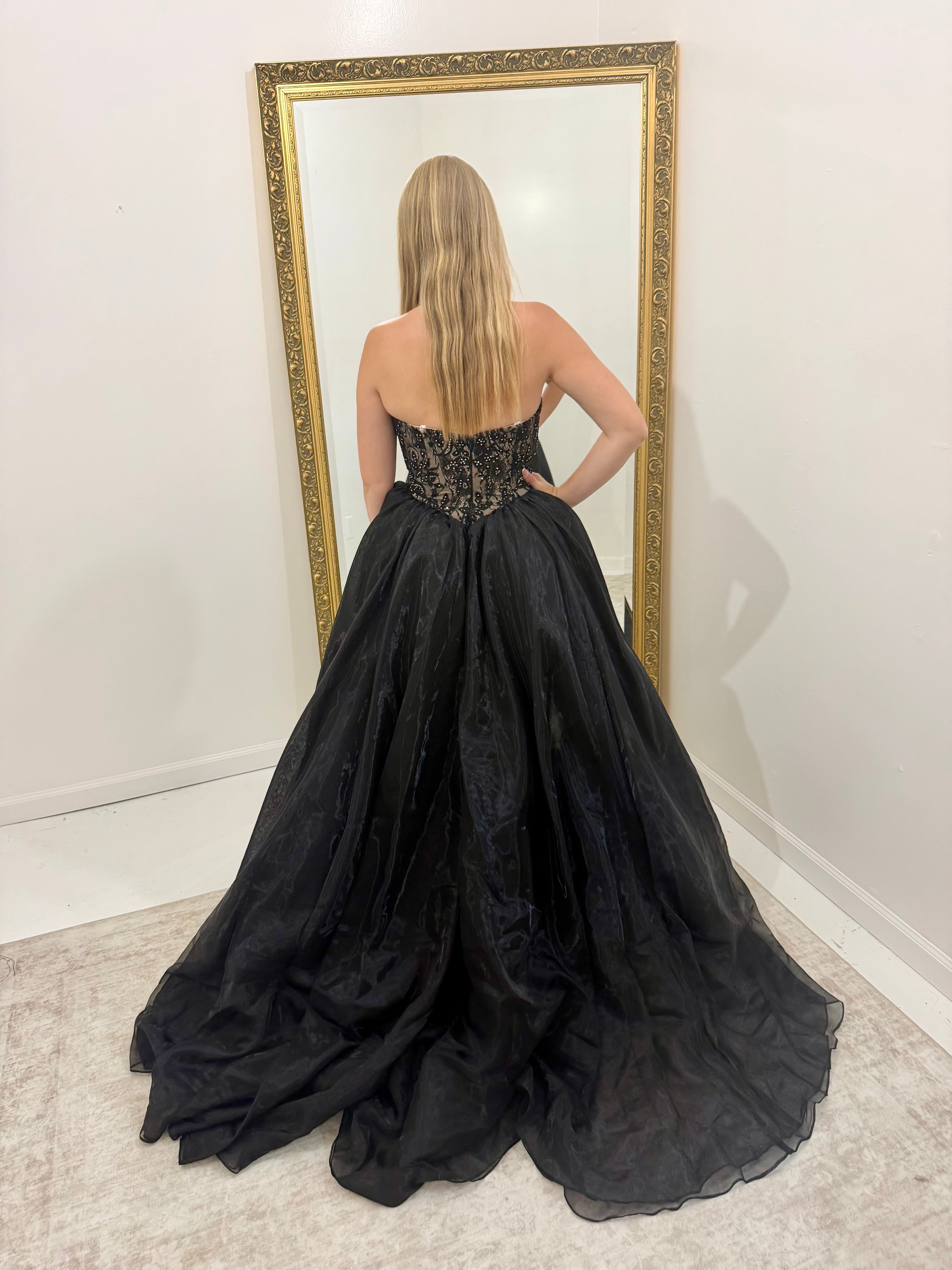 A woman stands with her back to the camera wearing a strapless black ball gown with a sheer lace bodice and a voluminous tulle skirt, posing in front of a large gold-framed mirror.