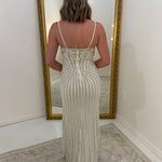 A woman stands with her back to the camera wearing a sleeveless, floor-length fitted dress with vertical pleated detailing and thin straps. She is positioned in front of a large gold-framed floor mirror in a room with white walls and a light rug.
