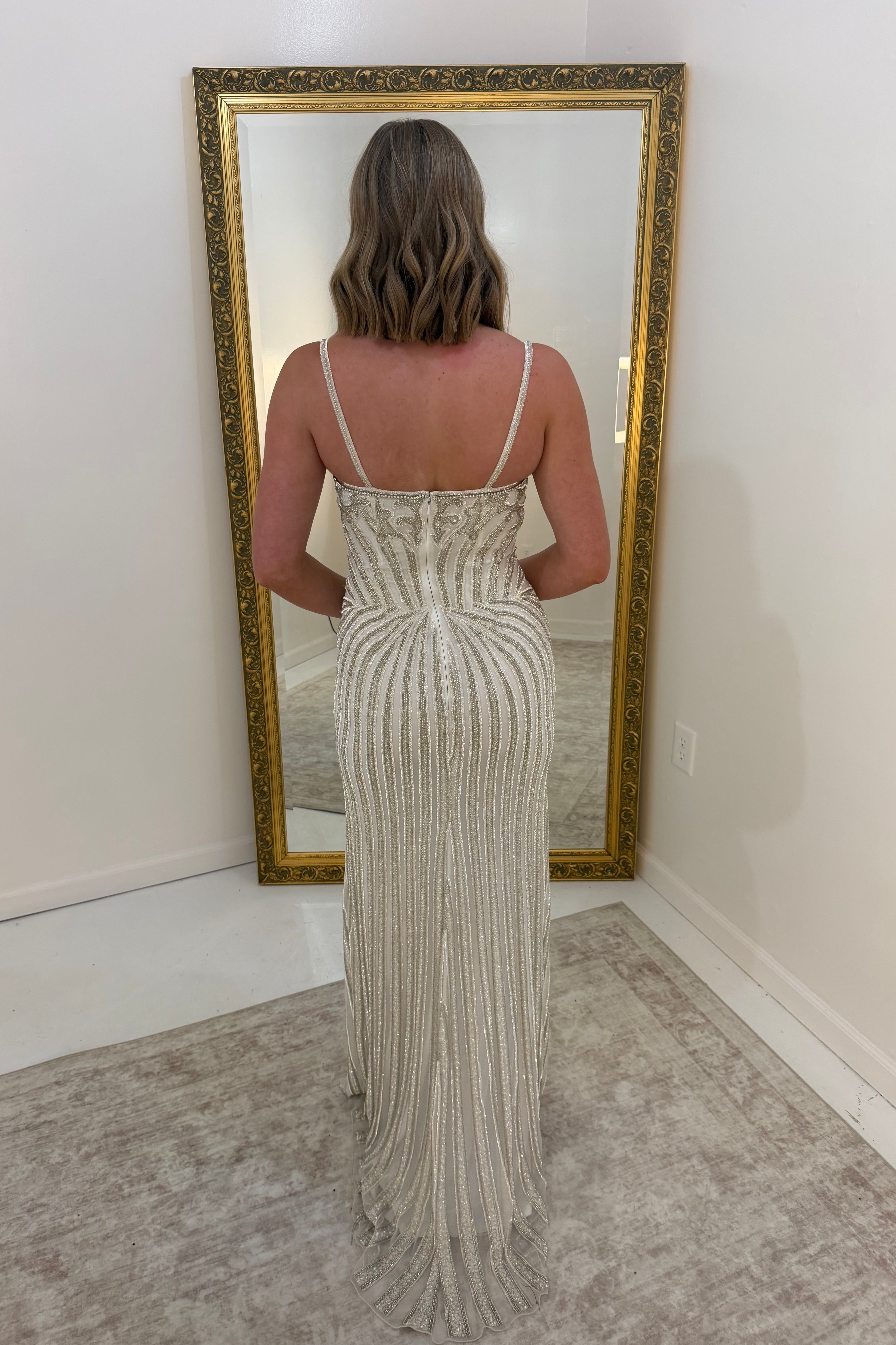 A woman stands with her back to the camera wearing a sleeveless, floor-length fitted dress with vertical pleated detailing and thin straps. She is positioned in front of a large gold-framed floor mirror in a room with white walls and a light rug.