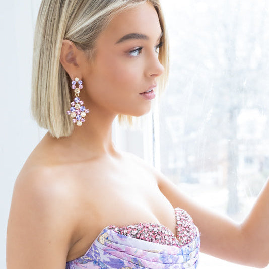 A woman wearing a strapless lavender and pink floral gown with a corseted bodice, posing by a bright window and wearing pink crystal drop earrings.