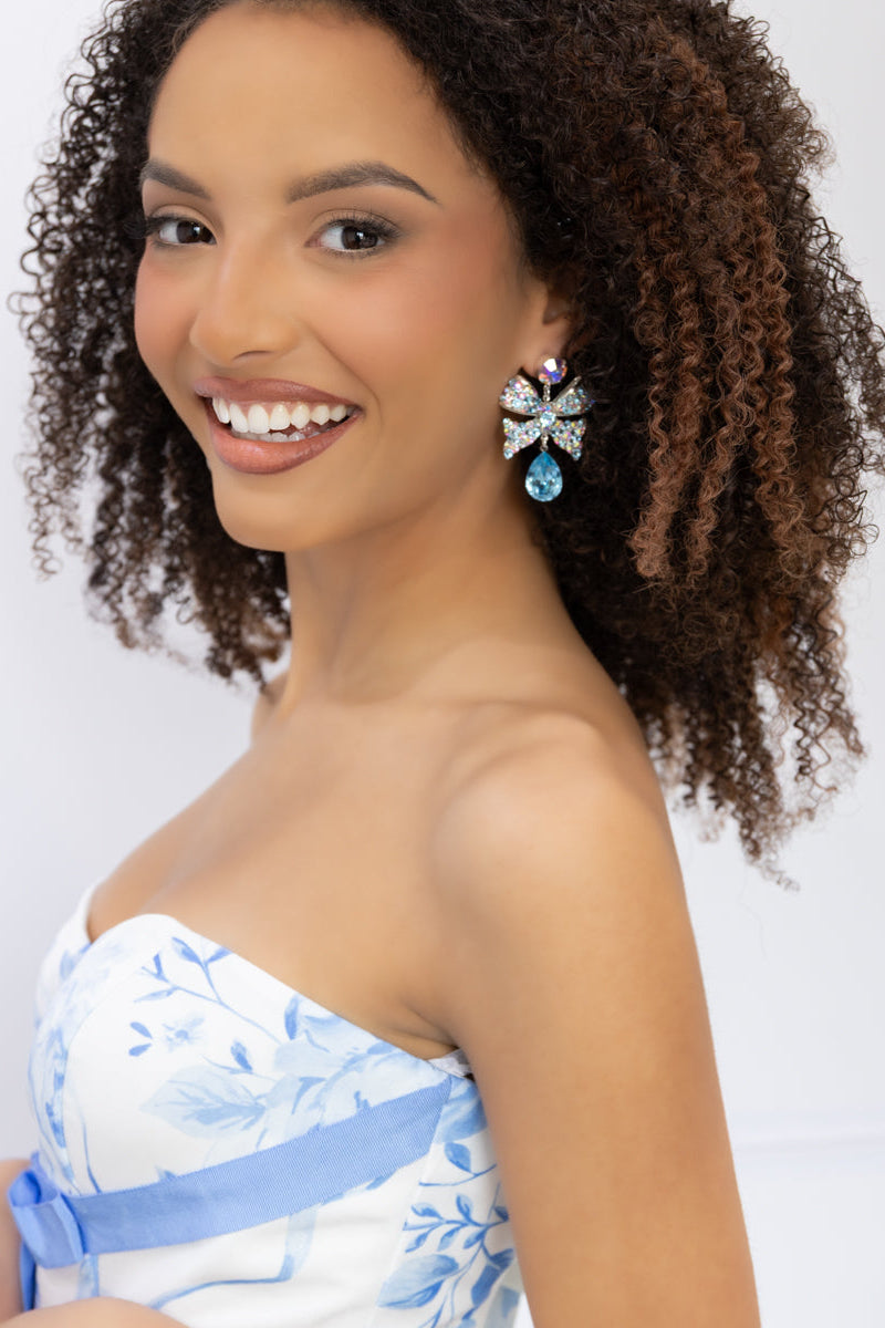 A smiling woman with curly brown hair wearing a strapless white dress with a light blue floral pattern and a blue sash, accessorized with a large blue and silver bow earring, against a plain white background.