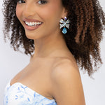 A smiling woman with curly brown hair wearing a strapless white dress with a light blue floral pattern and a blue sash, accessorized with a large blue and silver bow earring, against a plain white background.