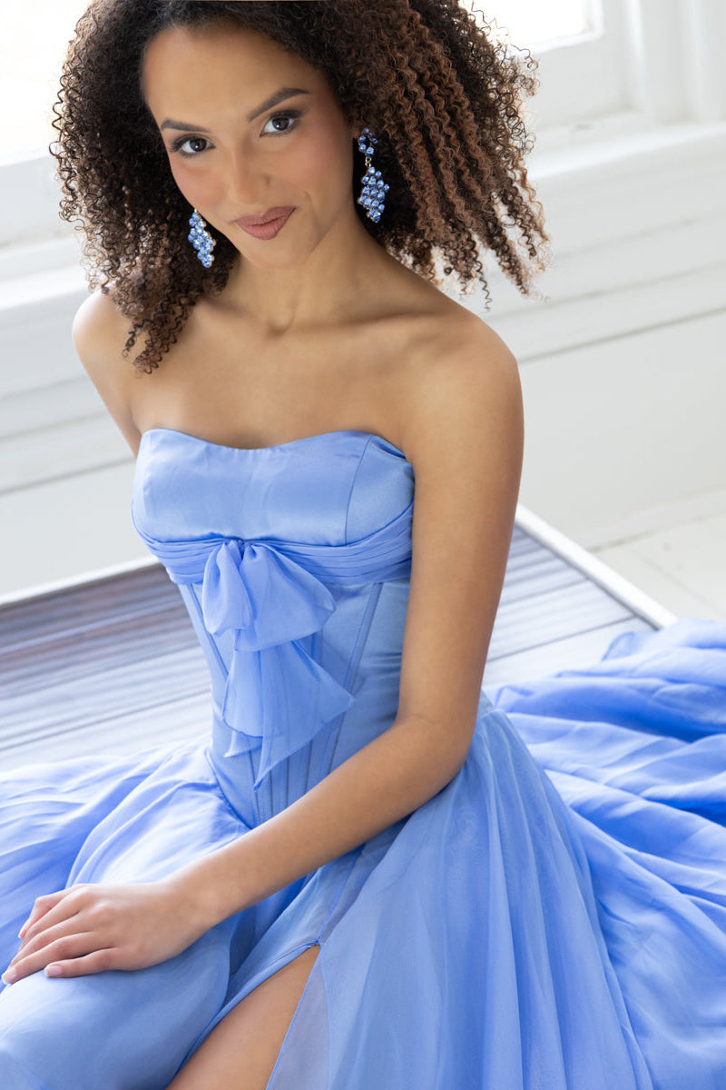 A woman wearing a strapless light blue formal gown with a large bow at the bust a flowing skirt, and matching light blue earrings. Seated indoors near a window.