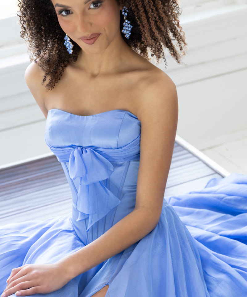 A woman wearing a strapless light blue formal gown with a large bow at the bust a flowing skirt, and matching light blue earrings. Seated indoors near a window.