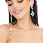 A close-up of a woman wearing a white floral gown and matching blue-and-white patterned earrings, against a plain white background.