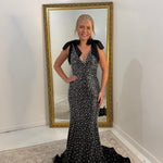 A woman stands indoors wearing a fitted black evening gown covered in small white polka dots, with short black sleeves and a deep V-neckline. She is posed in front of a tall floor mirror with a gold frame on a light rug against white walls.