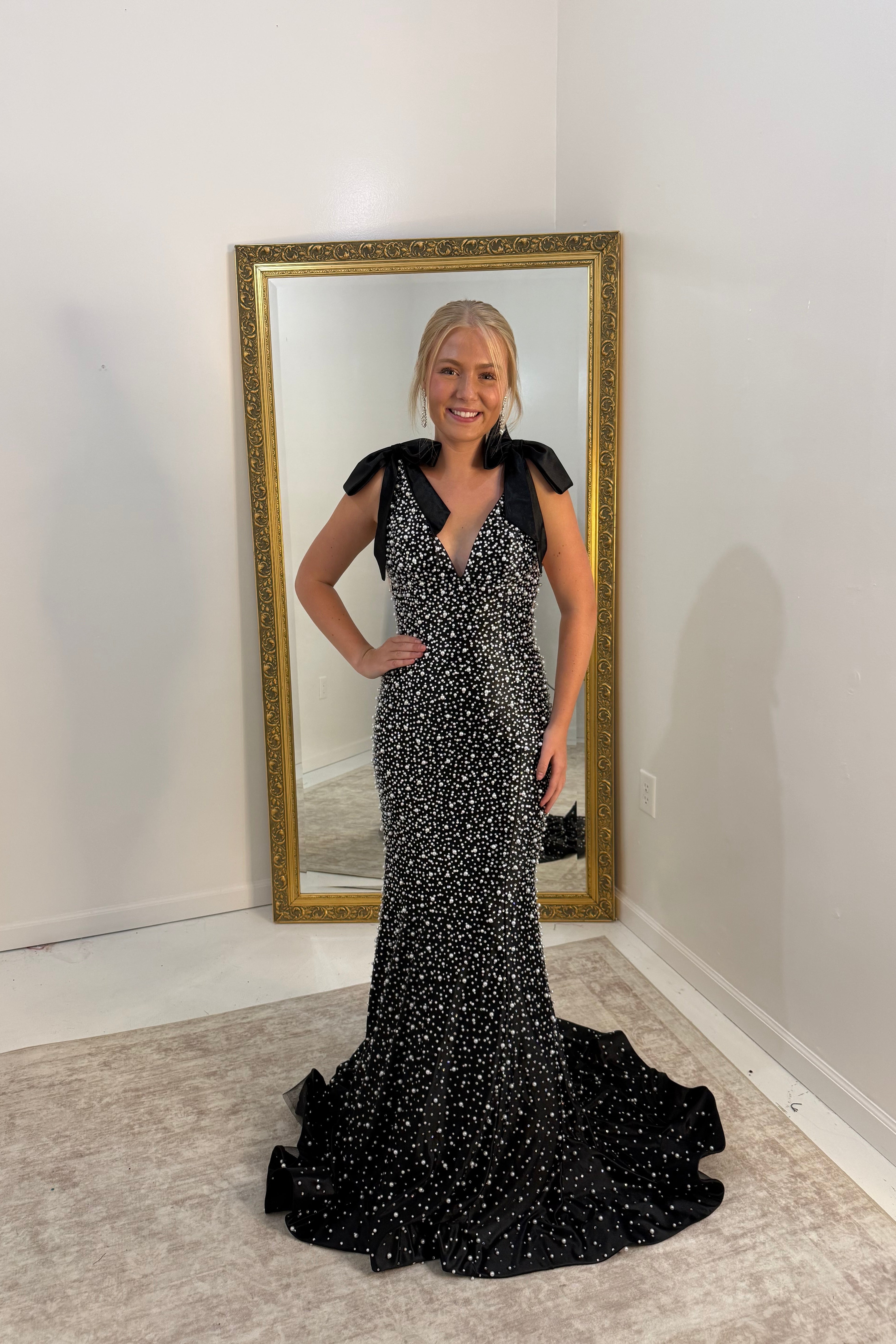 A woman stands indoors wearing a fitted black evening gown covered in small white polka dots, with short black sleeves and a deep V-neckline. She is posed in front of a tall floor mirror with a gold frame on a light rug against white walls.