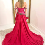 A woman stands with her back to the camera wearing a bright fuchsia pink off-the-shoulder ball gown with a long train, standing in front of a large gold-framed mirror.