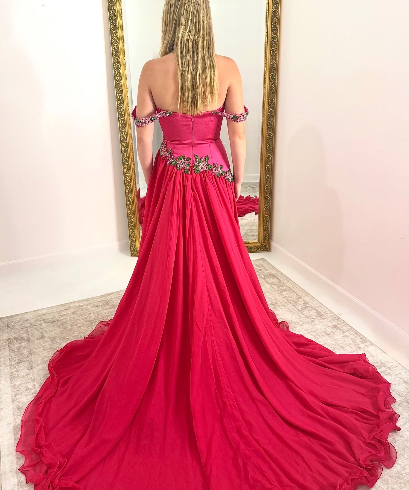 A woman stands with her back to the camera wearing a bright fuchsia pink off-the-shoulder ball gown with a long train, standing in front of a large gold-framed mirror.