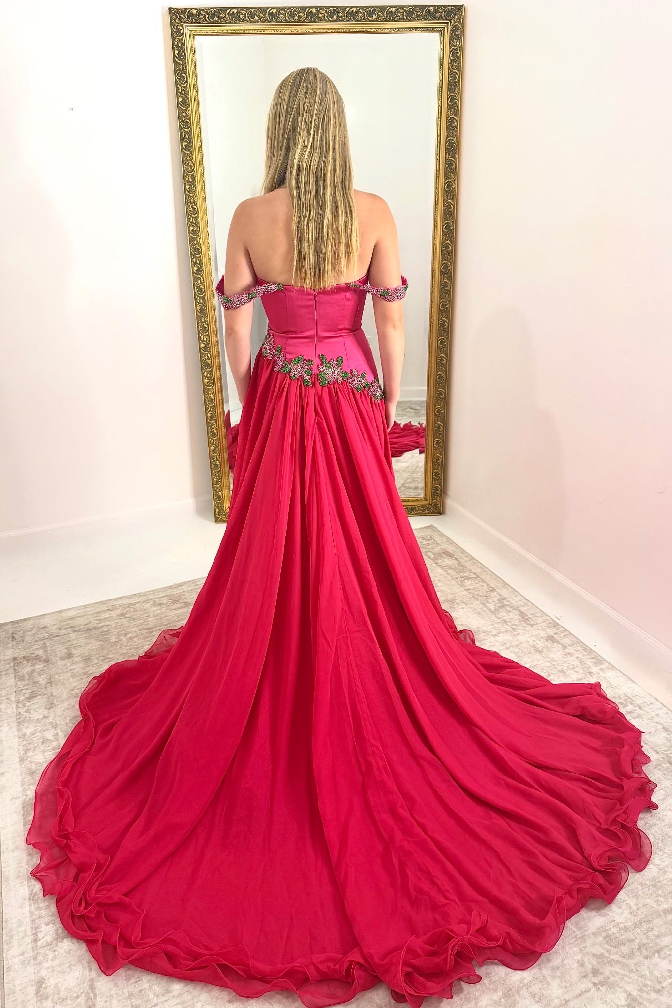 A woman stands with her back to the camera wearing a bright fuchsia pink off-the-shoulder ball gown with a long train, standing in front of a large gold-framed mirror.