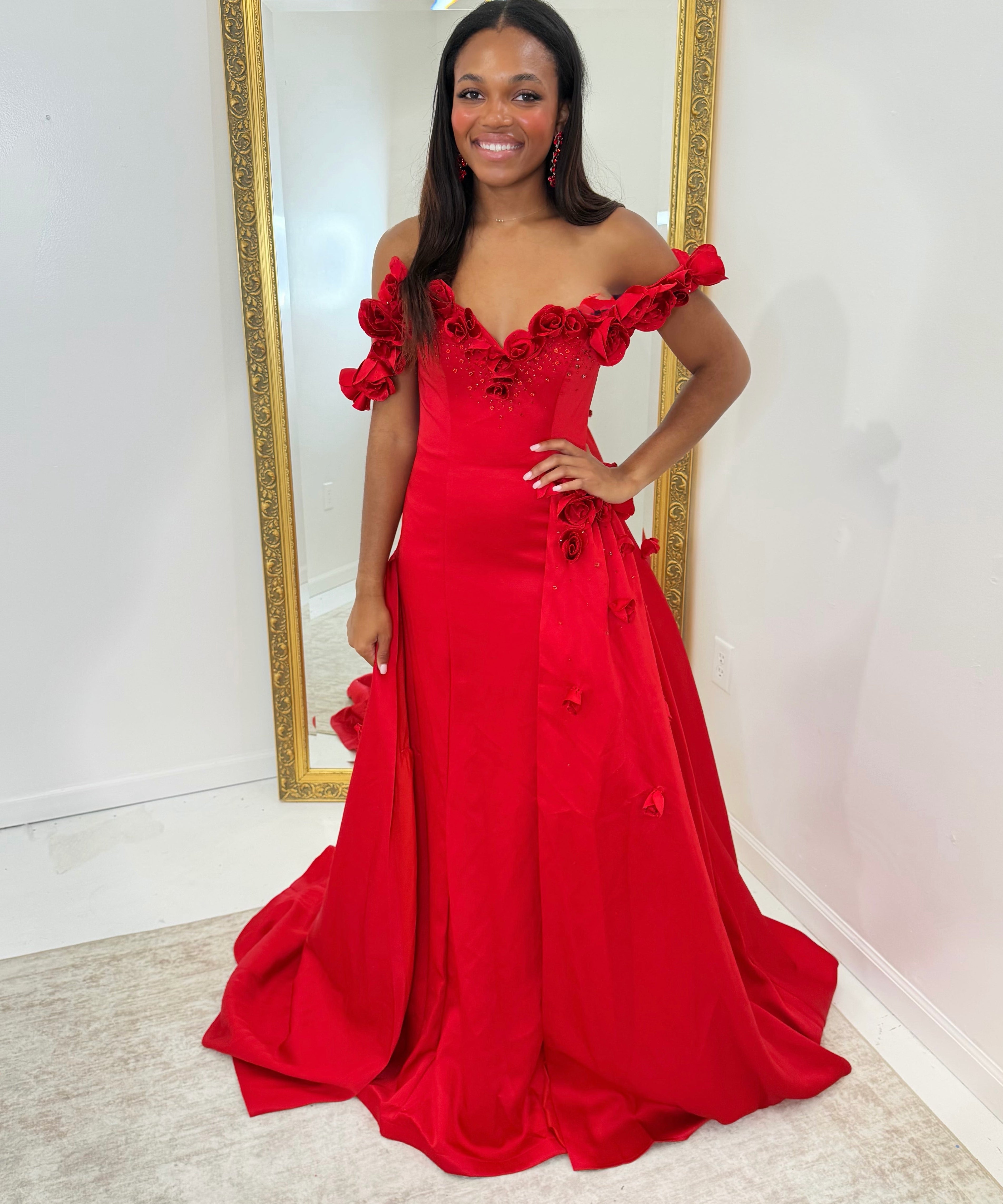 A woman wearing a bright red off-the-shoulder evening gown with 3D floral appliqué details, standing in front of a large gold-framed mirror in a white room.