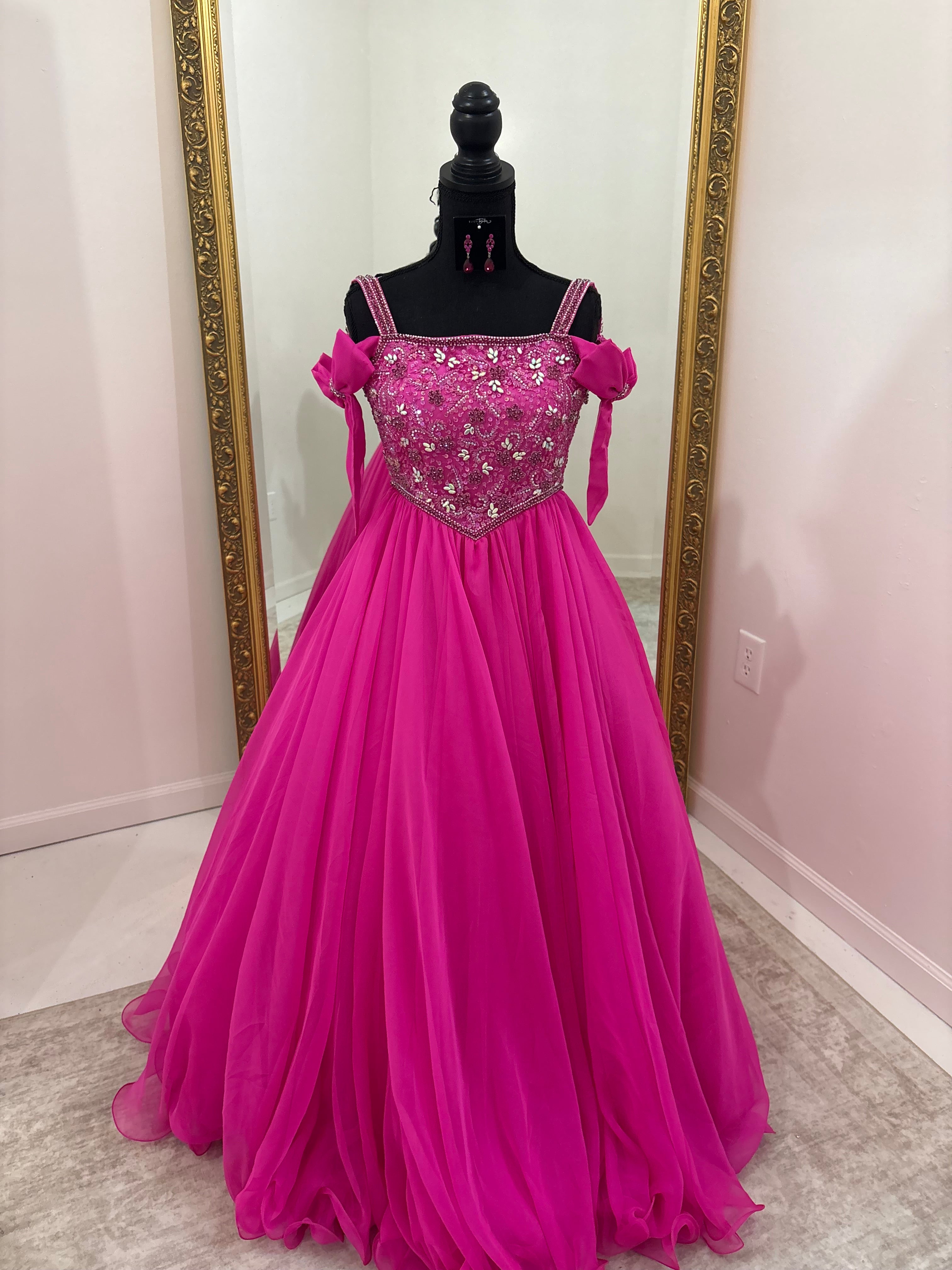 A bright fuchsia pink off-the-shoulder ball gown with a heavily embellished beaded bodice, displayed on a black mannequin in front of a mirror with a gold frame.