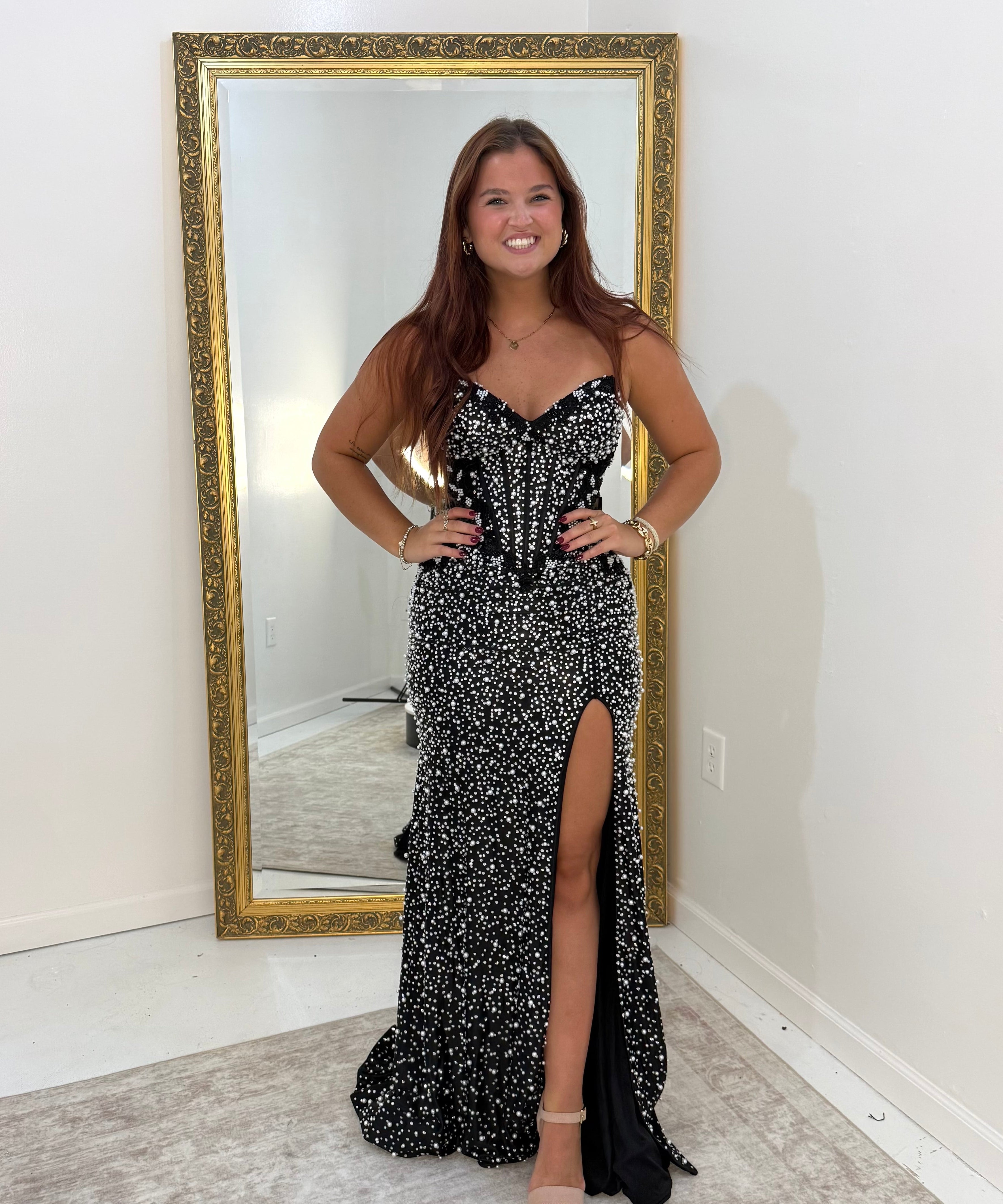 A woman stands in a room wearing a black and white beaded strapless gown with a sweetheart neckline, corset-style bodice, and a high thigh slit. She is posing in front of a large gold-framed mirror.