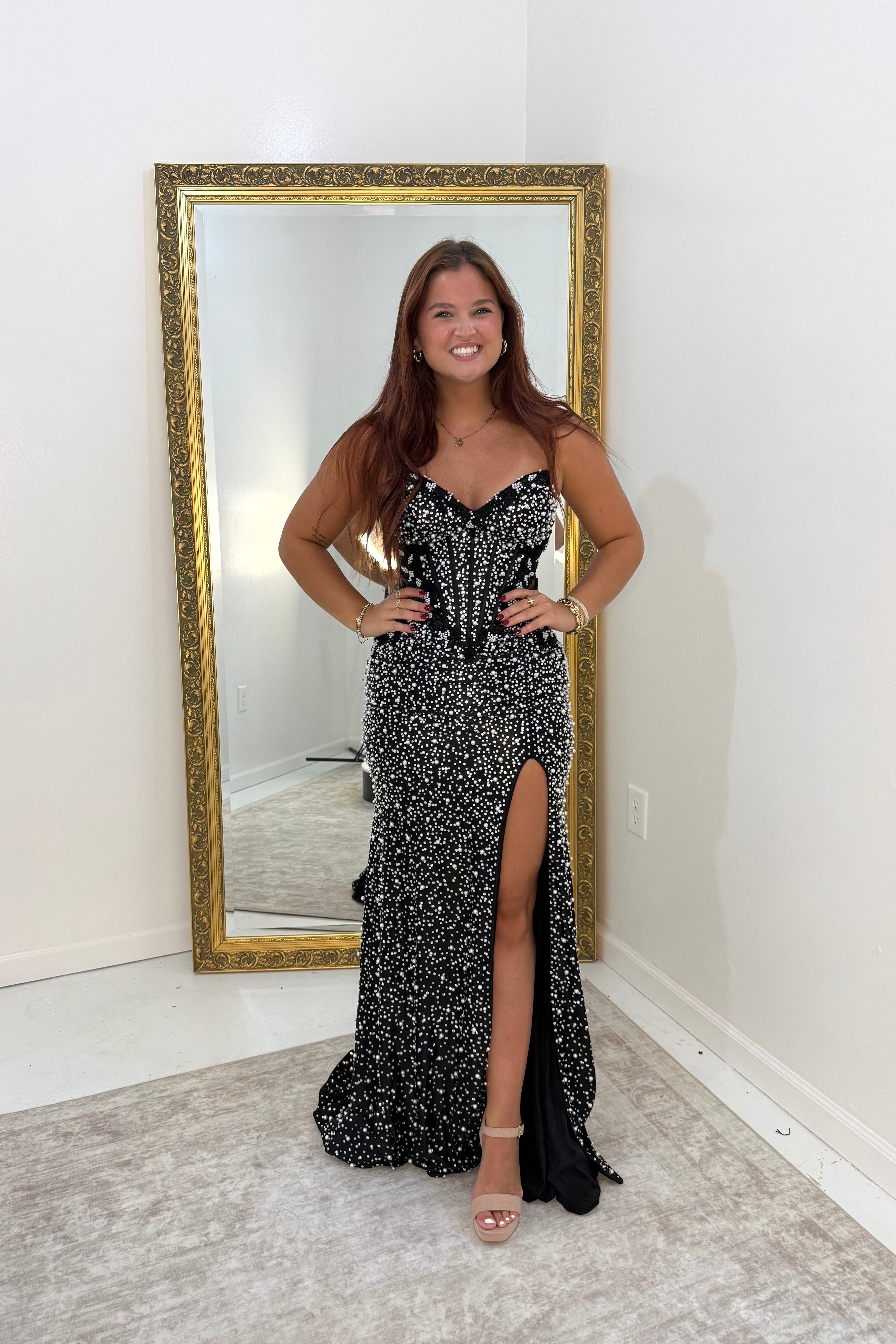 A woman stands in a room wearing a black and white beaded strapless gown with a sweetheart neckline, corset-style bodice, and a high thigh slit. She is posing in front of a large gold-framed mirror.