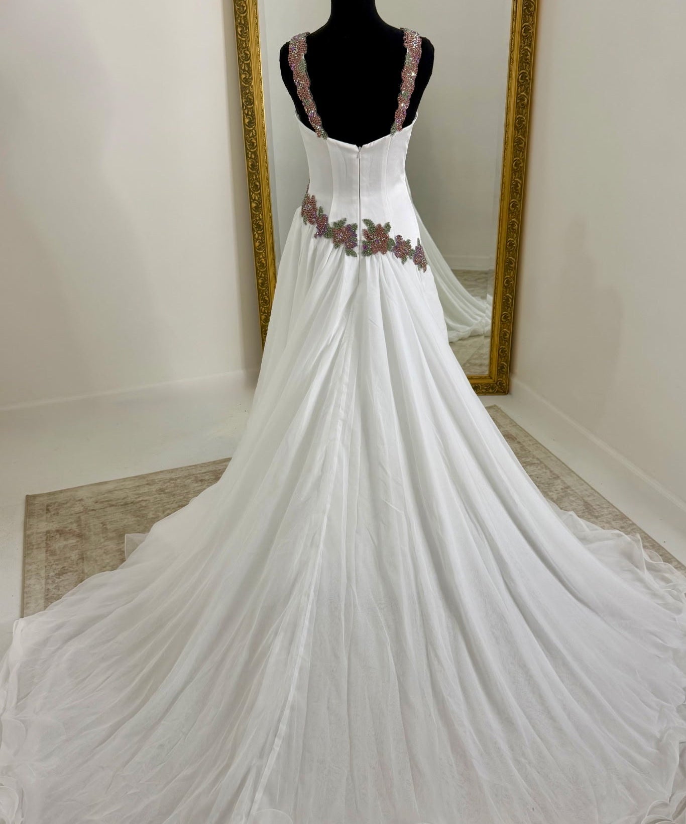 A white gown displayed on a black mannequin in front of a mirror, featuring a fitted bodice with floral embroidery at the waist and a long flowing train.