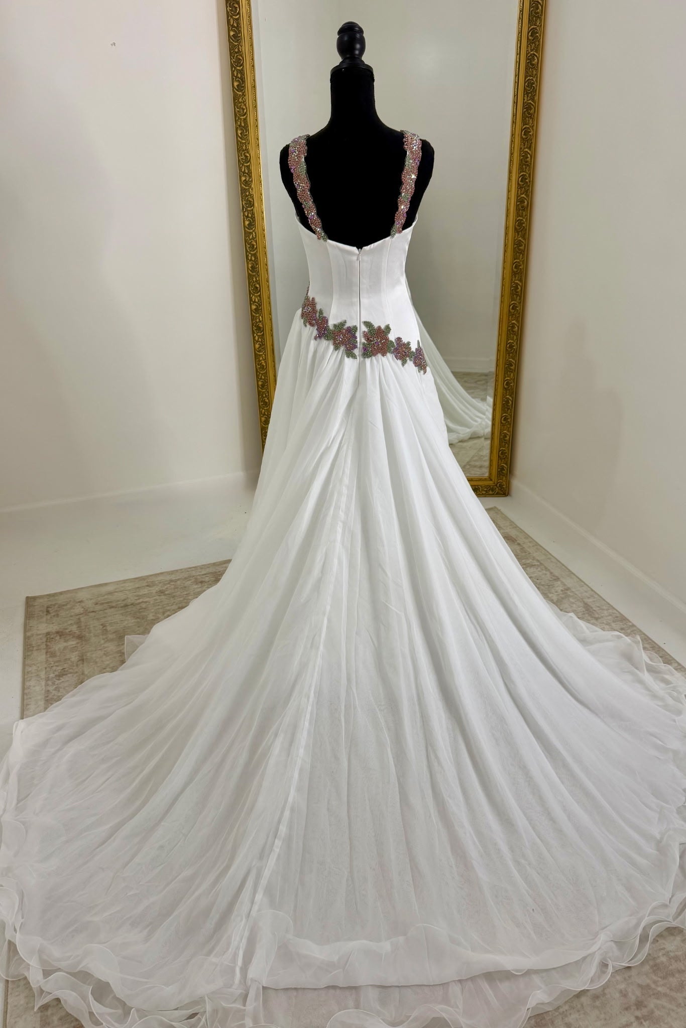 A white gown displayed on a black mannequin in front of a mirror, featuring a fitted bodice with floral embroidery at the waist and a long flowing train.