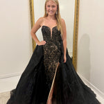 A woman modeling a strapless black lace formal gown with a sweetheart neckline, thigh-high slit, and a dramatic black tulle overskirt, standing in front of a gold-framed mirror.