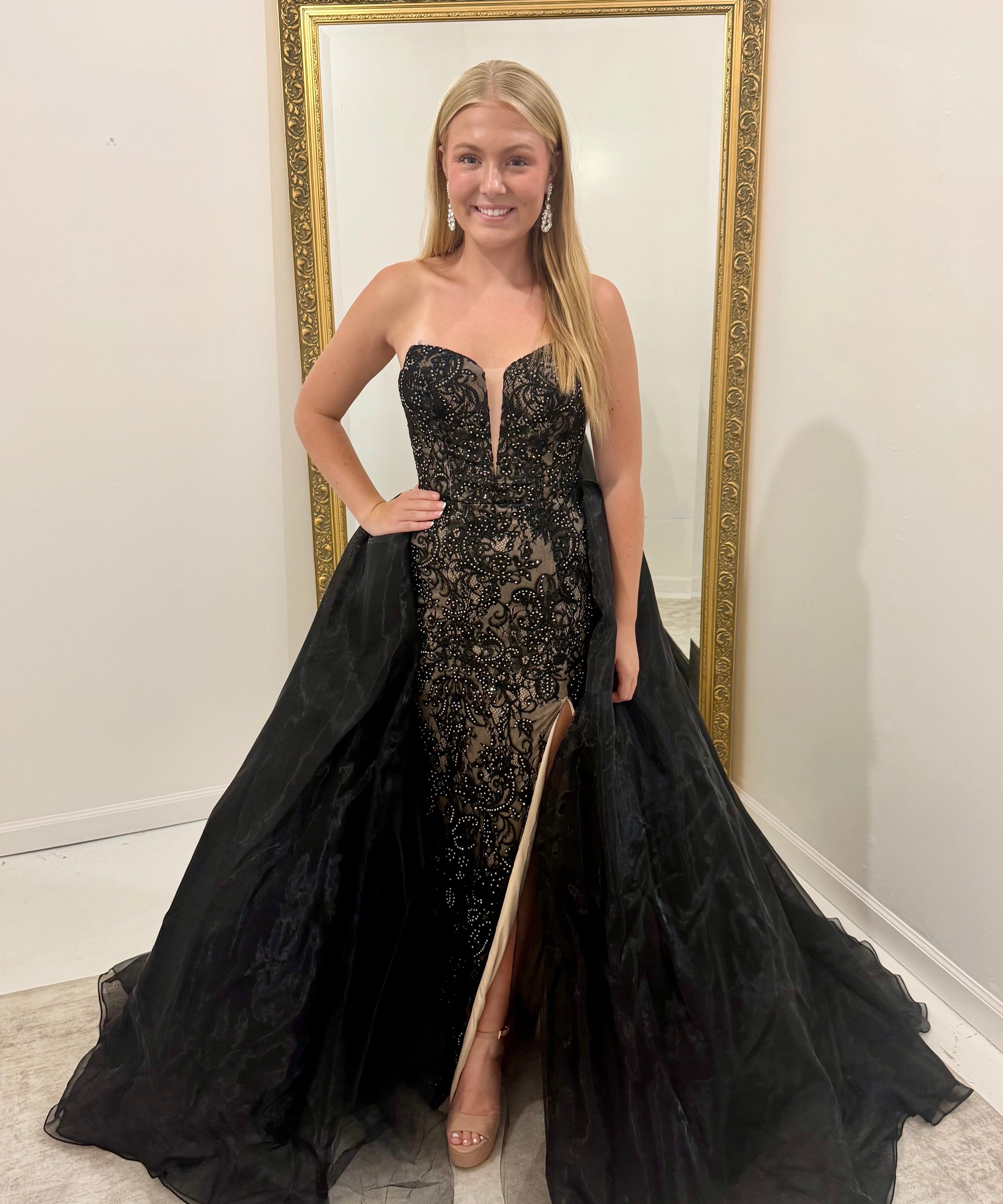A woman modeling a strapless black lace formal gown with a sweetheart neckline, thigh-high slit, and a dramatic black tulle overskirt, standing in front of a gold-framed mirror.